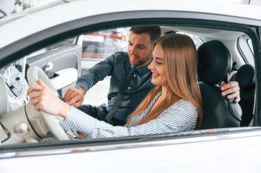 Otomobillerin nasıl çalıştığını anlatıyor. Resmi elbiseli adam elektrikli arabalı kadın müşteriyle birlikte..
