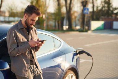 Sıkıldım, otomobilin şarj olmasını bekliyorum. Akıllı telefonlu. Adam dışarıda elektrikli arabasının yanında duruyor..