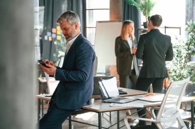 Adam masaya yaslanmış durumda. Akıllı telefon ellerindeyken. Bir grup iş adamı modern ofiste çalışıyor..