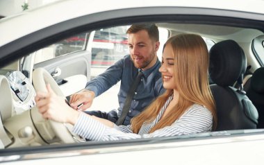 Otomobillerin nasıl çalıştığını anlatıyor. Resmi elbiseli adam elektrikli arabalı kadın müşteriyle birlikte..
