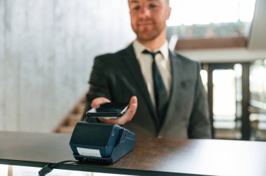 Ödemeyi telefon ve terminal kullanarak yapıyor. Otel resepsiyonunda siyah ceketli bir adam var..