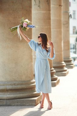 Beton sütunların yanında duruyordu. Güzel bir kadın elinde çiçeklerle dışarıda bekliyor..