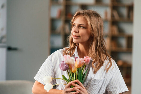 Holding the gift. Young woman is standing in domestic room with flowers.
