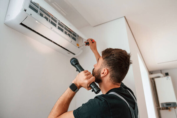 Using screwdriver and flashlight. Man repairs an air conditioner indoors at home.