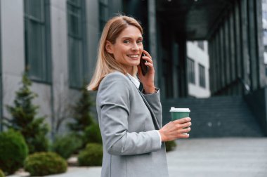 Yürümek, bir bardak içki içmek ve telefonla konuşmak. Resmi giysili kadın iş binasının yakınında dışarıda..