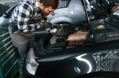 Tamirci kaputun altındaki arabayı tamir etmek için bir araç kullanıyor. Üniformalı adam araba galerisinde çalışıyor..