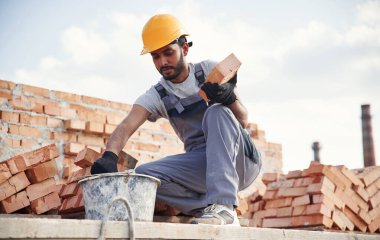 Bucket on the ground, holding a brick. Handsome Indian man is on the construction site. clipart