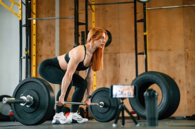 Yan görünüş, halterli. Tripodun üzerindeki akıllı telefon video kaydediyor. Güzel, güçlü kadın spor salonunda..