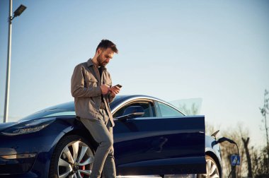 Otomobili şarj ediyorum. Adam dışarıda elektrikli arabasının yanında duruyor..