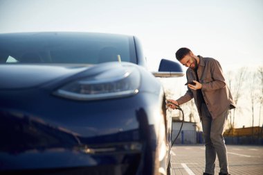 Yandan bak. Otomobili şarj ediyorum. Adam dışarıda elektrikli arabasının yanında duruyor..