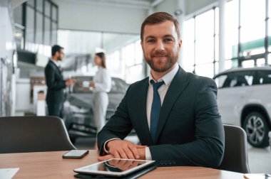 Kulaklıklı adam masanın yanında oturuyor. Araba galerisinde üç kişi birlikte çalışıyor..