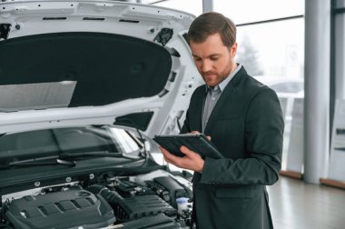 Ayakta, yandan, tableti tutarak. Siyah takım elbiseli adam otomobilin yanındaki araba galerisinde..