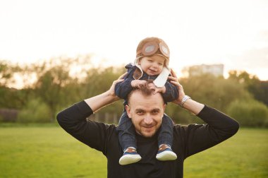 Çocuk insanın omuzlarında oturuyor. Baba ve küçük oğlu sahada oyuncak uçakla oynuyorlar..