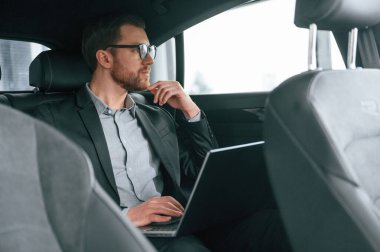 Gözlükle, dizüstü bilgisayarla çalışarak. Resmi iş elbiseli adam modern otomobilde oturuyor..