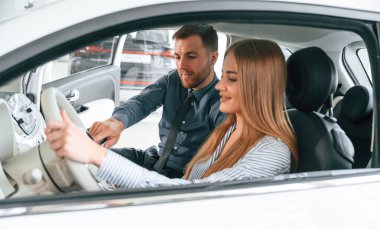 Otomobillerin nasıl çalıştığını anlatıyor. Resmi elbiseli adam elektrikli arabalı kadın müşteriyle birlikte..
