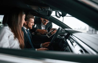 Yan görüş. Araba galerisi çalışanı, otomobilin içinde oturan kadın ve erkeğe danışmanlık yapıyor..