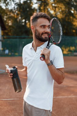 Su ve raketle şişeyi tutuyor. Genç adam güneşli bir günde tenis kortunda..