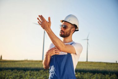 Mavi üniformalı ve güneş gözlüklü. Servis mühendisi yel değirmenleriyle sahada..