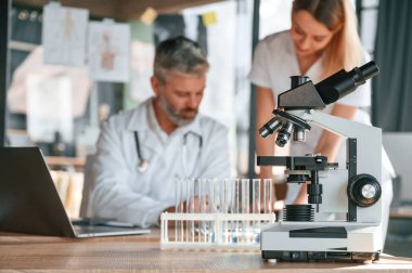 Laptop, test tüpleri, mikroskop. İki klinik çalışanı içeride birlikte. Genç bir kadınla kıdemli bir adam.