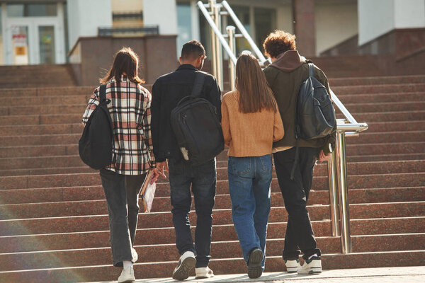 View from back, walking together. Four young students in casual clothes are outdoors.