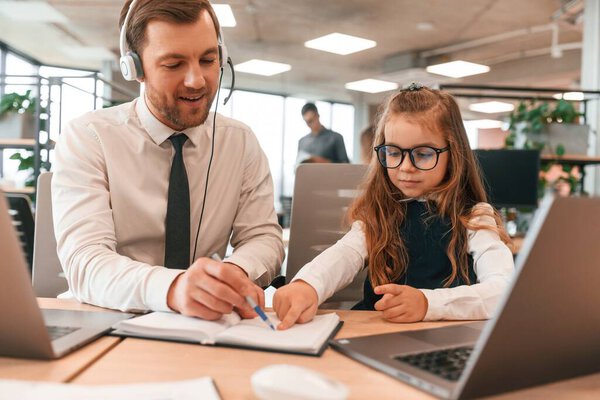 Man is helping, teaching. Little girl is in the office where people working together.