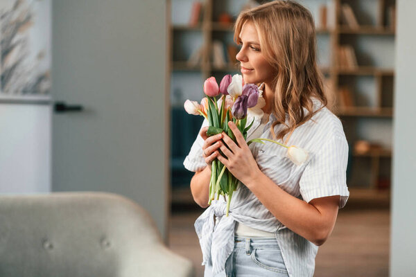 Positive young woman is standing in domestic room with flowers.