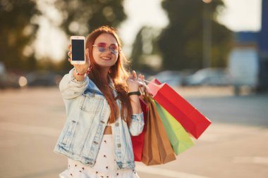 Elindeki akıllı telefonu gösteriyordun. Günlük giysiler içinde güzel bir kadın alışveriş torbaları tutuyor, açık havada..