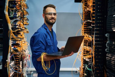 Ellerinde dizüstü bilgisayarla. Genç adam sunucu odasında internet ekipmanı ve kablolarıyla çalışıyor..