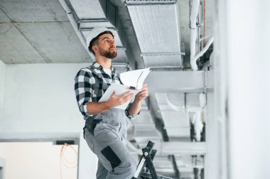 Evraklar ellerinde. Beyaz kasketli. Üniformalı tamirci tamamlanmamış binada..