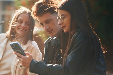 Kız akıllı telefondan bir şey gösteriyor. Üniversitenin dışında üç genç öğrenci var..