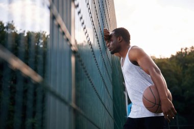 Metal yapıya yaslanarak. Genç siyah adam basketbol topuyla dışarıda..