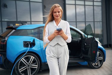 Akıllı telefon kullanıyor. Elektrikli arabalardaki kadın gündüz şarj istasyonunda.