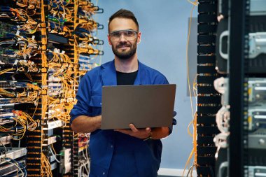 Ellerinde dizüstü bilgisayarla. Genç adam sunucu odasında internet ekipmanı ve kablolarıyla çalışıyor..