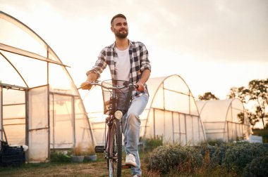 Ön manzara. Günlük giysiler içinde yakışıklı bir adam seranın yanındaki tarımsal alanda bisikletle geziyor..
