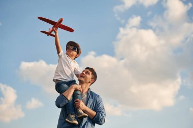 Turuncu renkli oyuncak uçak ellerinde. Baba ve küçük oğlu dışarıda oynuyor ve eğleniyorlar..