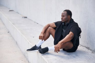 Beyaz duvarın yanında oturuyor. Yakışıklı siyah adam gündüz vakti dışarıda poz veriyor..