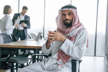 Ciddi bir yüz ifadesi. Geleneksel kıyafetli Müslüman işadamı ve görevde meslektaşları.