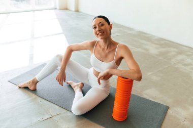 Köpüklü rulo ile oturuyor. Zayıf vücutlu genç bir kadın stüdyoda spor kıyafetleri giyiyor..