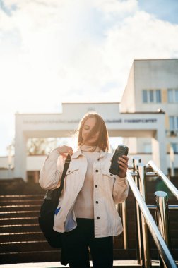 Güneş ışığıyla aydınlatılmış, elinde bir şişe su var. Gündelik giysiler içinde genç bir kız öğrenci dışarıda..
