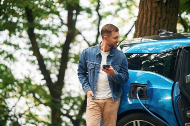 Ayakta ve akıllı telefonu tutarak. Yakışıklı adam dışarıda elektrikli arabasının şarj olmasını bekliyor..