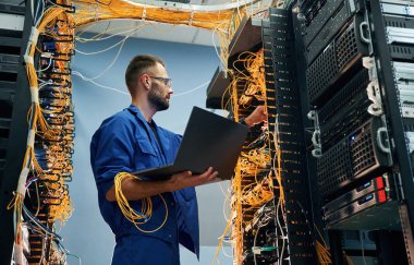 Ellerinde dizüstü bilgisayarla. Genç adam sunucu odasında internet ekipmanı ve kablolarıyla çalışıyor..