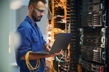 Gümüş renkli dizüstü bilgisayar ellerinde. Genç adam sunucu odasında internet ekipmanı ve kablolarıyla çalışıyor..