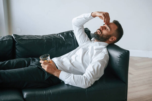 Голова болит, лежит на диване. Расстроенный молодой человек пьет алкоголь в помещении.
