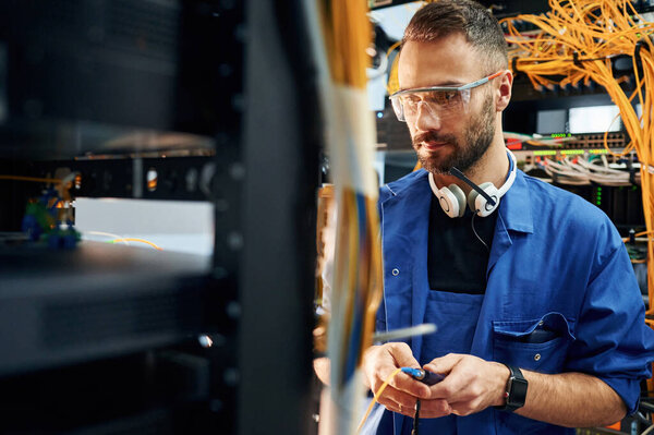 В форме синего цвета. Молодой человек работает с интернет-оборудованием и проводами в серверной.