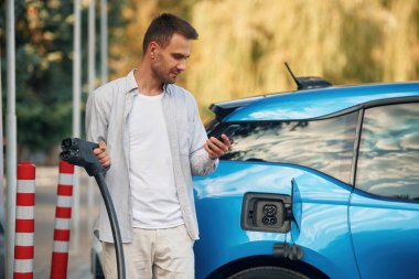 Prizde kablo var, akıllı telefon tutuyor. Şarj istasyonunda mavi elektrikli arabası olan adam..