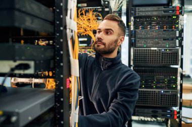 Siyah kıyafetler. Genç adam sunucu odasında internet ekipmanı ve kablolarıyla çalışıyor..