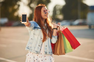 Elindeki akıllı telefonu gösteriyordun. Günlük giysiler içinde güzel bir kadın alışveriş torbaları tutuyor, açık havada..