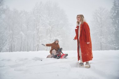 Bir kadın kızak çekiyor. Anne ve kızı kış çayırlarında ve ormandalar..