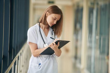 Elinde not defteriyle. Beyaz önlüklü kadın doktor koridorda..