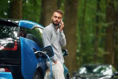 Kablosuz cihaz, akıllı telefon kullanıyor. Yakışıklı adam dışarıda elektrikli otomobilin yanında duruyor ve şarj edilmeyi bekliyor..